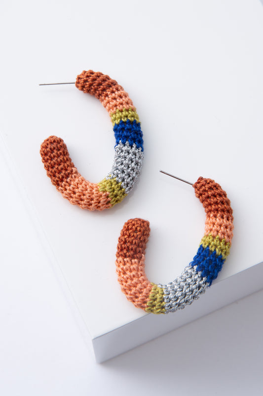 The Maza Crocheted Hoops are oval-shaped hoops covered entirely in crocheted thread. The thread color alternates between stripes of silver, mustard, peach, rust, and blue. The alternating stripes vary in width and create a color-blocked look.