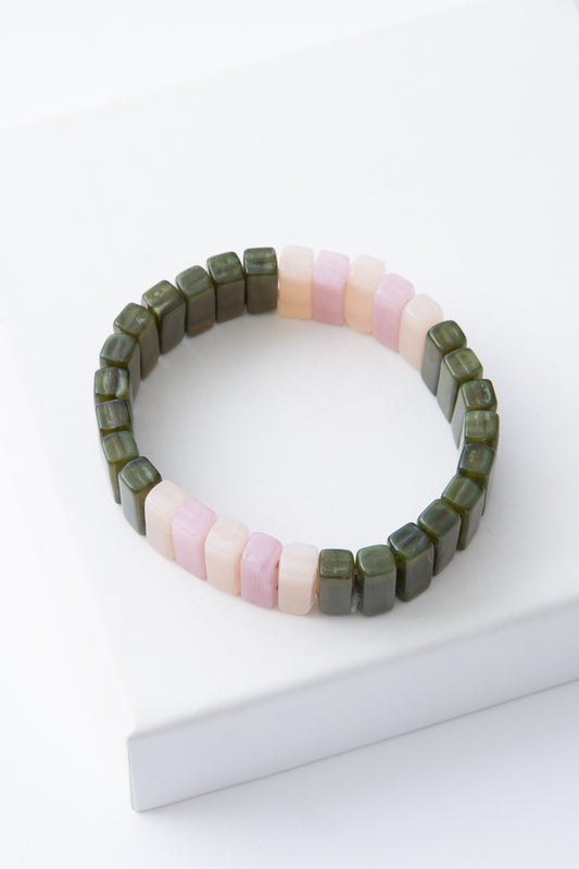 The Meadow Bracelet is a stretchy bracelet composed of rectangular glass beads strung on elastic. The beads alternate between olive sections and blush and white sections for a color-blocked look.