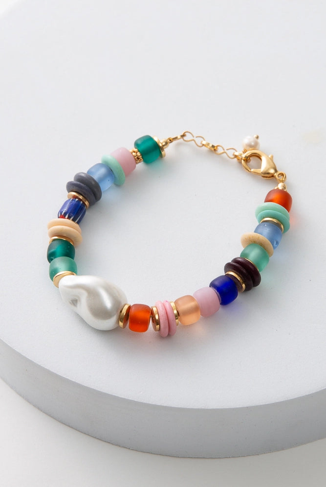 The Menagerie Bracelet is a single strand bracelet composed of a rainbow of glass beads. The beads include shades of red, pink, orange, blue, and green. Disc-shaped brass and bone beads are interspersed with the glass beads. A glass bead in the shape of a freshwater pearl sits at the front of the bracelet. The bracelet has a gold clasp.