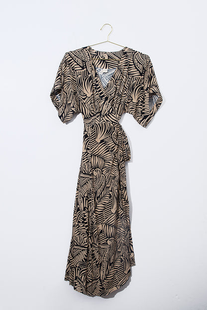 The Midi Wrap Dress in Mongabay hangs against a wall. It is a long, flowy dress made of a soft rayon blend fabric. The fabric has a tan background with black block-print inspired patterns covering it. The patterns have a tropical feel reminiscent of fruits and palm fronds. The dress has wide, short sleeves and hits the lower to mid calf. It has a v-neck and a fabric tie that wraps around the waist.