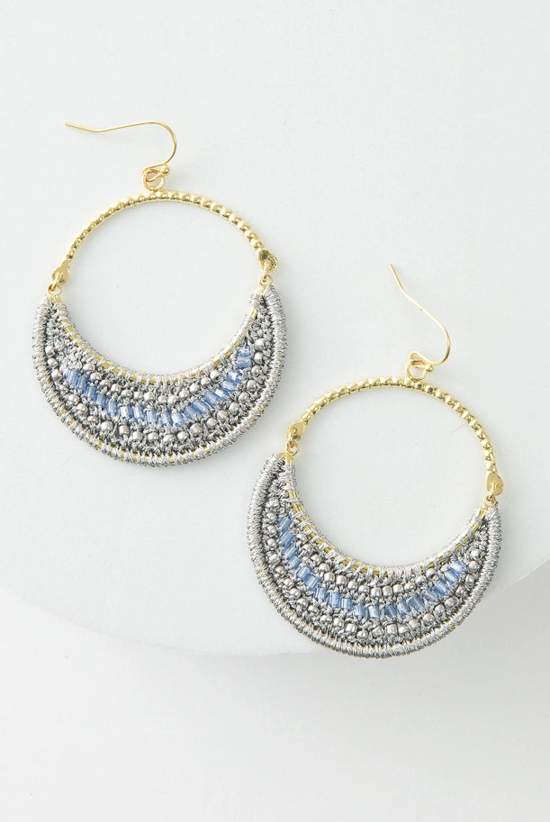 The Moonlight Earrings are hoop earrings. The top half of the hoop is gold metal. The bottom half of the hoop is filled with a crescent of silver thread and light blue glass beads.
