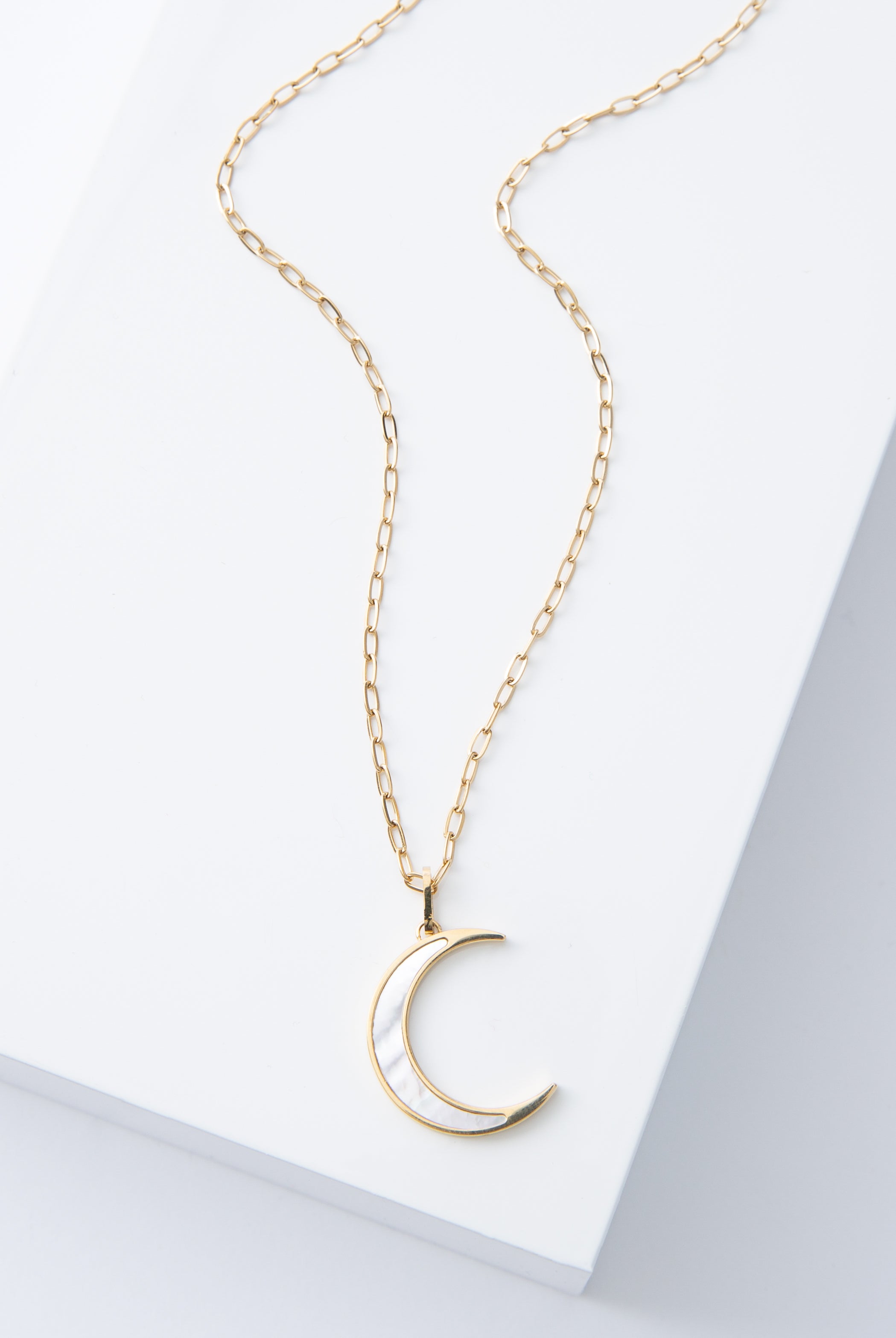 The Moonstruck Necklace is a gold chain necklace with a crescent moon shaped pendant. The pendant has a sharp, minimalist look. It is made of mother-of-pearl outlined in shining gold.