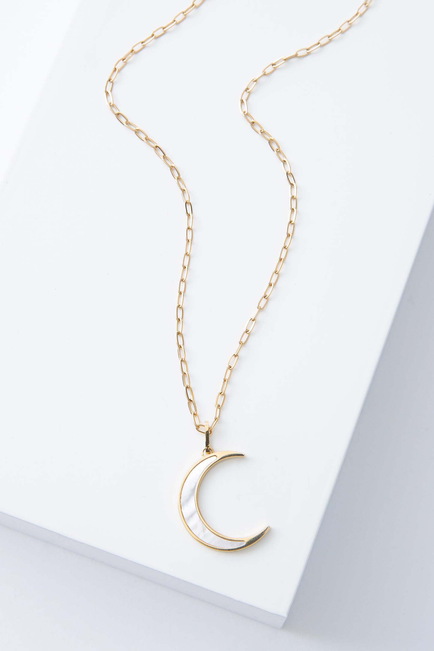 The Moonstruck Necklace is a gold chain necklace with a crescent moon shaped pendant. The pendant has a sharp, minimalist look. It is made of mother-of-pearl outlined in shining gold. 