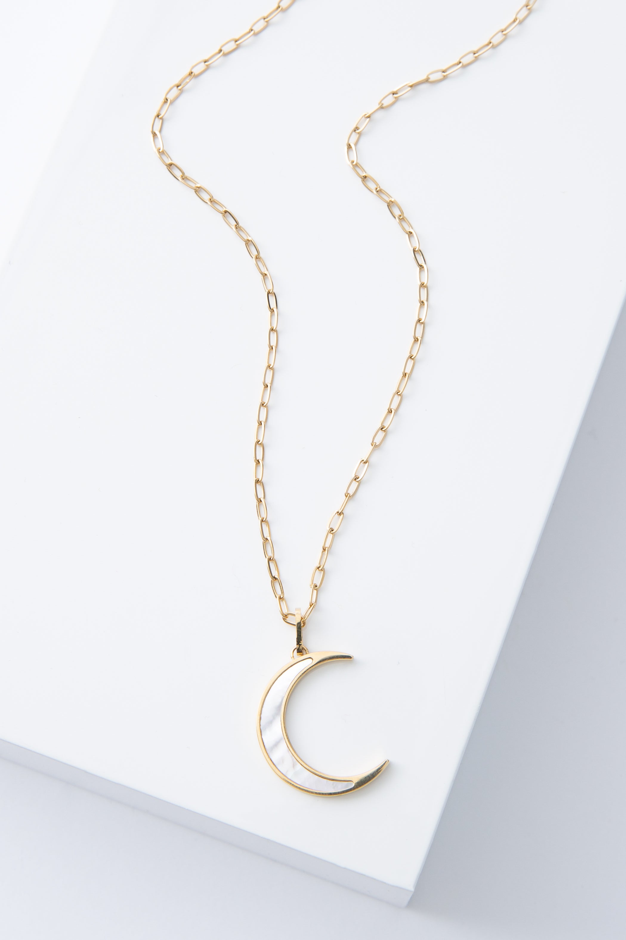 The Moonstruck Necklace is a gold chain necklace with a crescent moon shaped pendant. The pendant has a sharp, minimalist look. It is made of mother-of-pearl outlined in shining gold.
