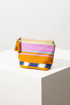 The Wander Pouch is a small, roughly square shaped pouch composed of striped cotton fabric. The stripes are gold, pink, blue, mint, and yellow, and vary in width. The pouch has a zip closure and there is a gold tassel attached to the zipper.