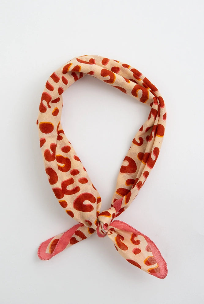 The On the Spot Bandana is a cotton bandana with a leopard print pattern. The background of the print is tan and the leopard spots are red. The bandana has a pink border.