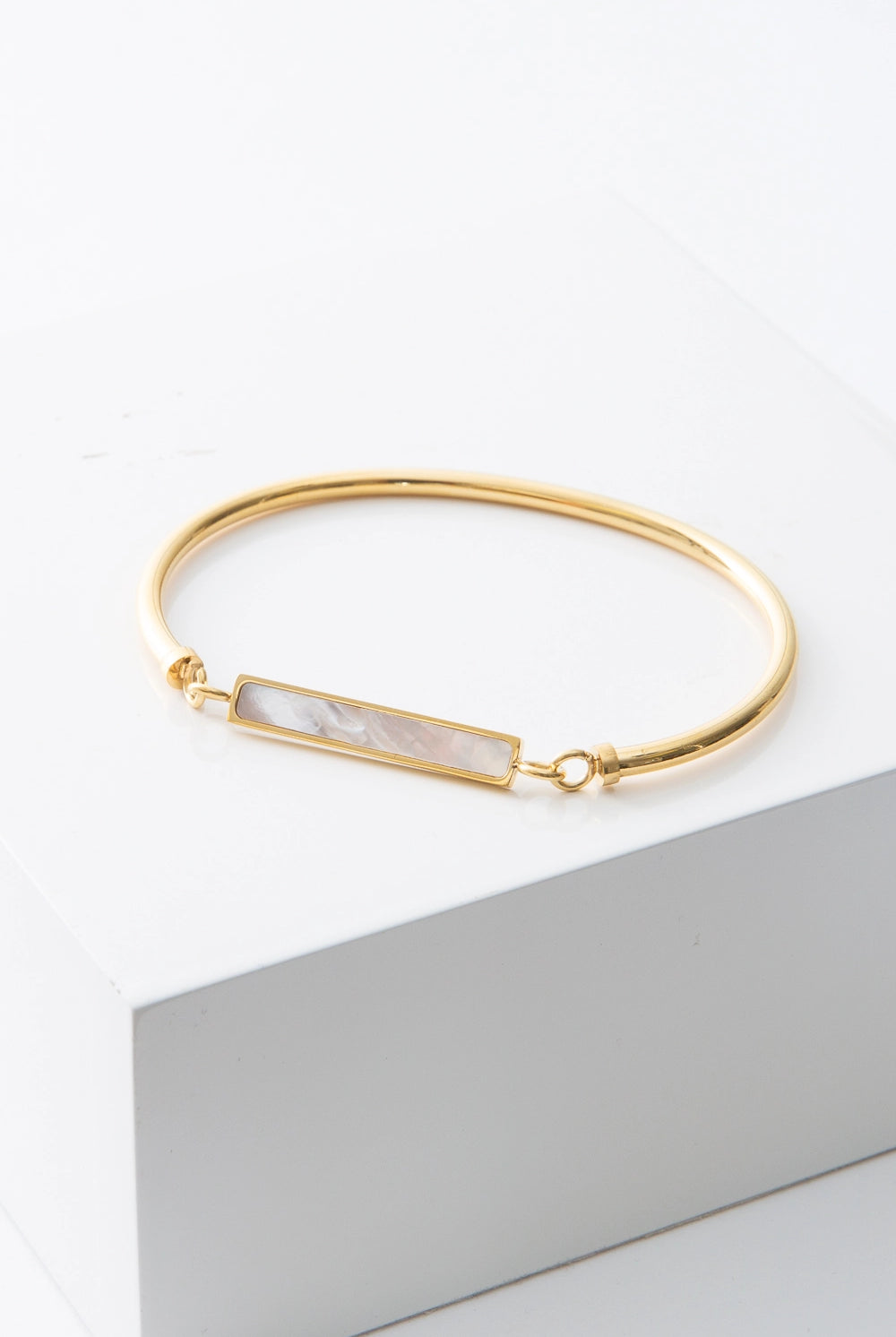 This is a thin cuff composed of one solid piece of gold-plated stainless steel that connects on either side to a narrow rectangular pendant that is suspended at the front of the cuff. The pendant is composed of shining mother-of-pearl outlined in gleaming gold.