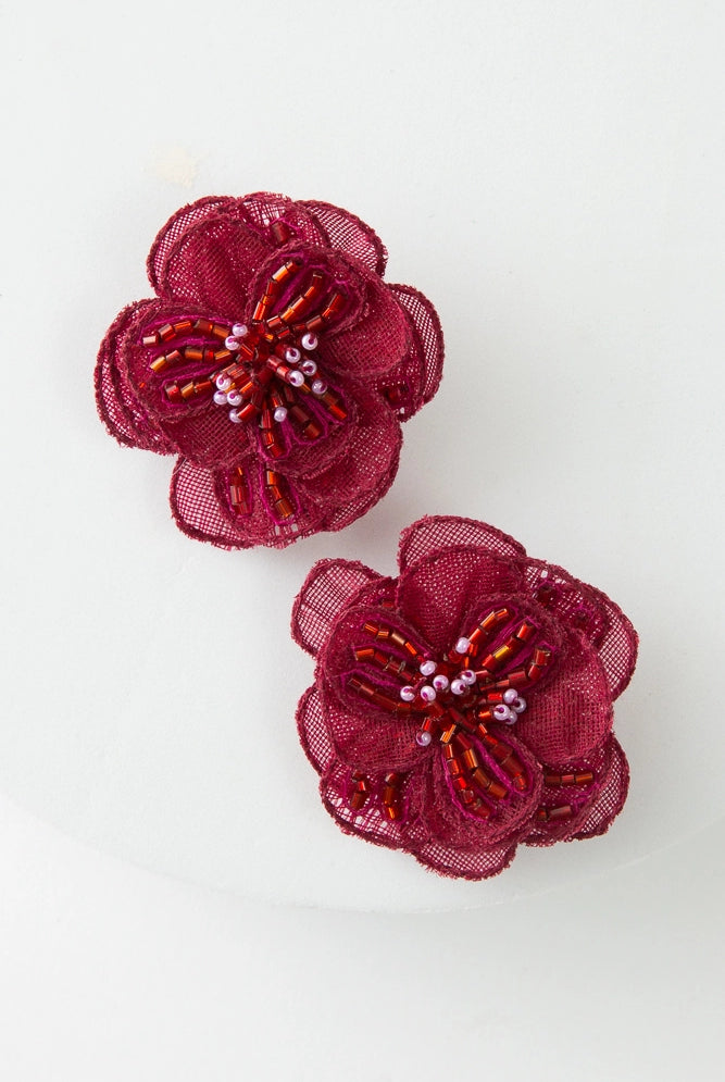 The Organza Earrings are large stud-style earrings composed of layers of crimson organza fabric petals that form a flower. In the middle of the flower are white and red glass beads that form the flower's center.