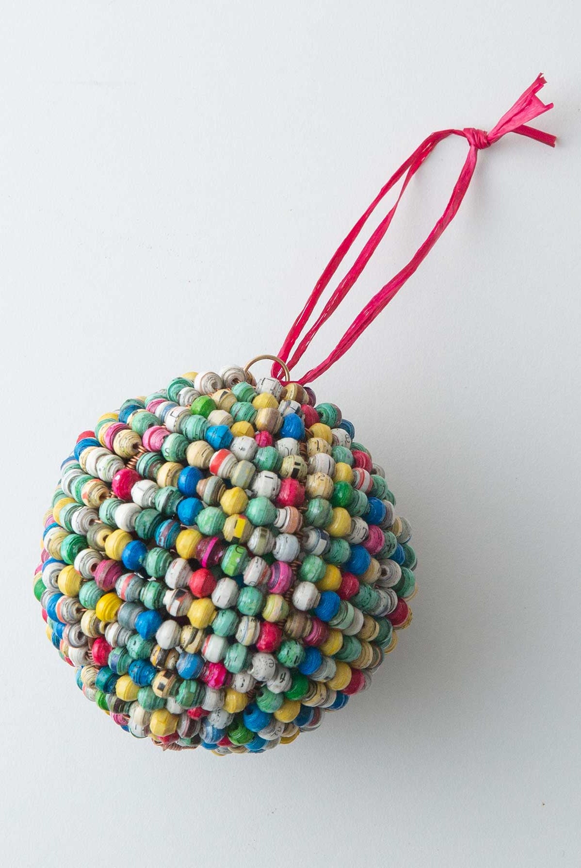 The Paper Sphere Ornament is a sphere shaped ornament composed entirely of small handrolled paper beads. The beads are every color of the rainbow. At the top of the ornament there is a red raffia loop that allows it to be hung.