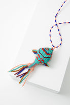 The Pescado Bag Charm is a three-dimensional beaded fish charm. The base of the fish is turquoise glass beads, and the fish's features are depicted using red, blue, pink, and purple beads. There is a beaded fringe at the fish's tail. The charm is attached to a beaded loop that allows it to be attached to a bag or other item.