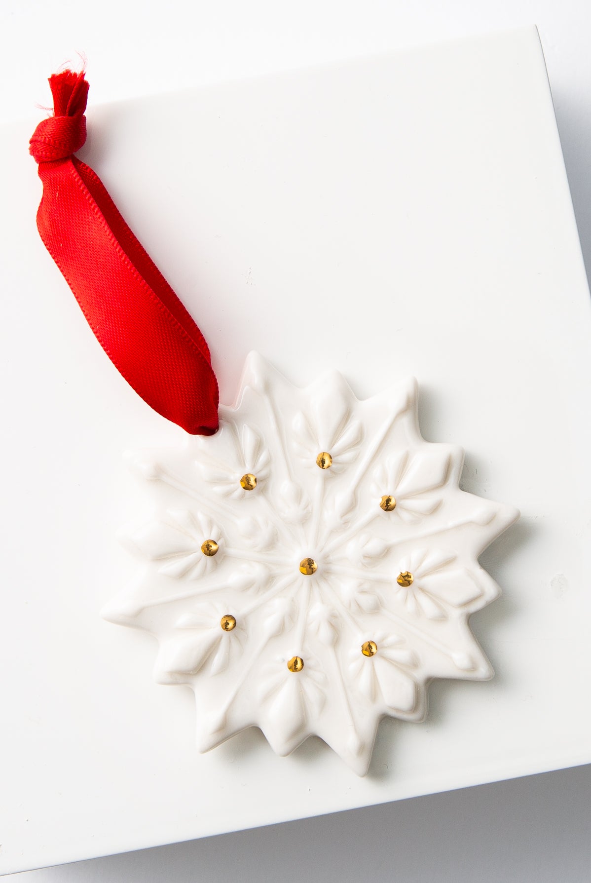The Porcelain Snowflake Ornament lays against a white background. It is a flat snowflake-shaped ornament made of shiny white porcelain. Gold dots accent the different points of the snowflake. A silky red ribbon attached to the top allows the ornament to be hung.
