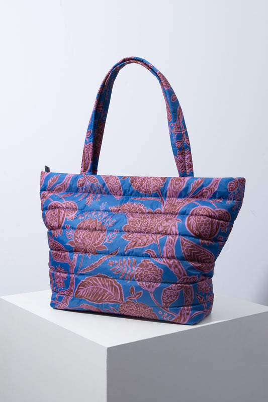 The Quilted Cotswold Tote is a large, square shaped tote that tapers up towards the top, making it slightly wider at the top than at the base. It is made of a quilted print fabric. The fabric has a blue background and a maroon and pink floral pattern. There is a zip closure at the top and two handles, allowing the bag to be slung over the shoulder. 