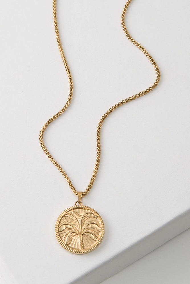 The Recharge Medallion is a gold pendant necklace. A round, gold pendant hangs from a gold chain. The pendant is etched with the image of a palm tree rising above a sand dune. The border of the pendant is etched with tiny lines, giving it a coin-like look.