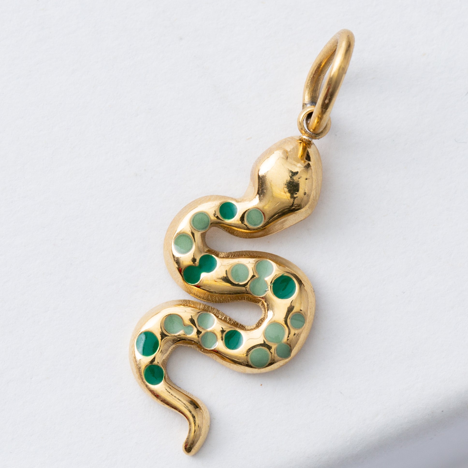 The Renew Charm features a winding gold snake charm attached to a brass loop. The snake is studded with light and dark green enamel dots along the length of its body.