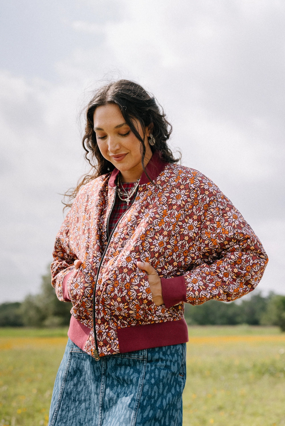 This reversible bomber jacket is composed of 100% cotton fabric. The fabric features a burgundy background with a white, yellow, and orange floral print with a mosaic-like look. The cuffs, collar, and bottom of the jacket are made of solid burgundy fabric. The bomber jacket has a zip closure up the center and two pockets.