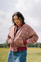 This reversible bomber jacket is composed of 100% cotton fabric. The fabric features a burgundy background with a white, yellow, and orange floral print with a mosaic-like look. The cuffs, collar, and bottom of the jacket are made of solid burgundy fabric. The bomber jacket has a zip closure up the center and two pockets.