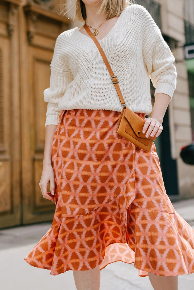 The Ruffle Wrap Skirt in Munni Sunset is a mid-length wrap skirt composed of a flowy rayon-cotton blend fabric. The fabric features an orange and pink tie-die style geometric print. The skirt hits just below the knee and has a ruffled hem.