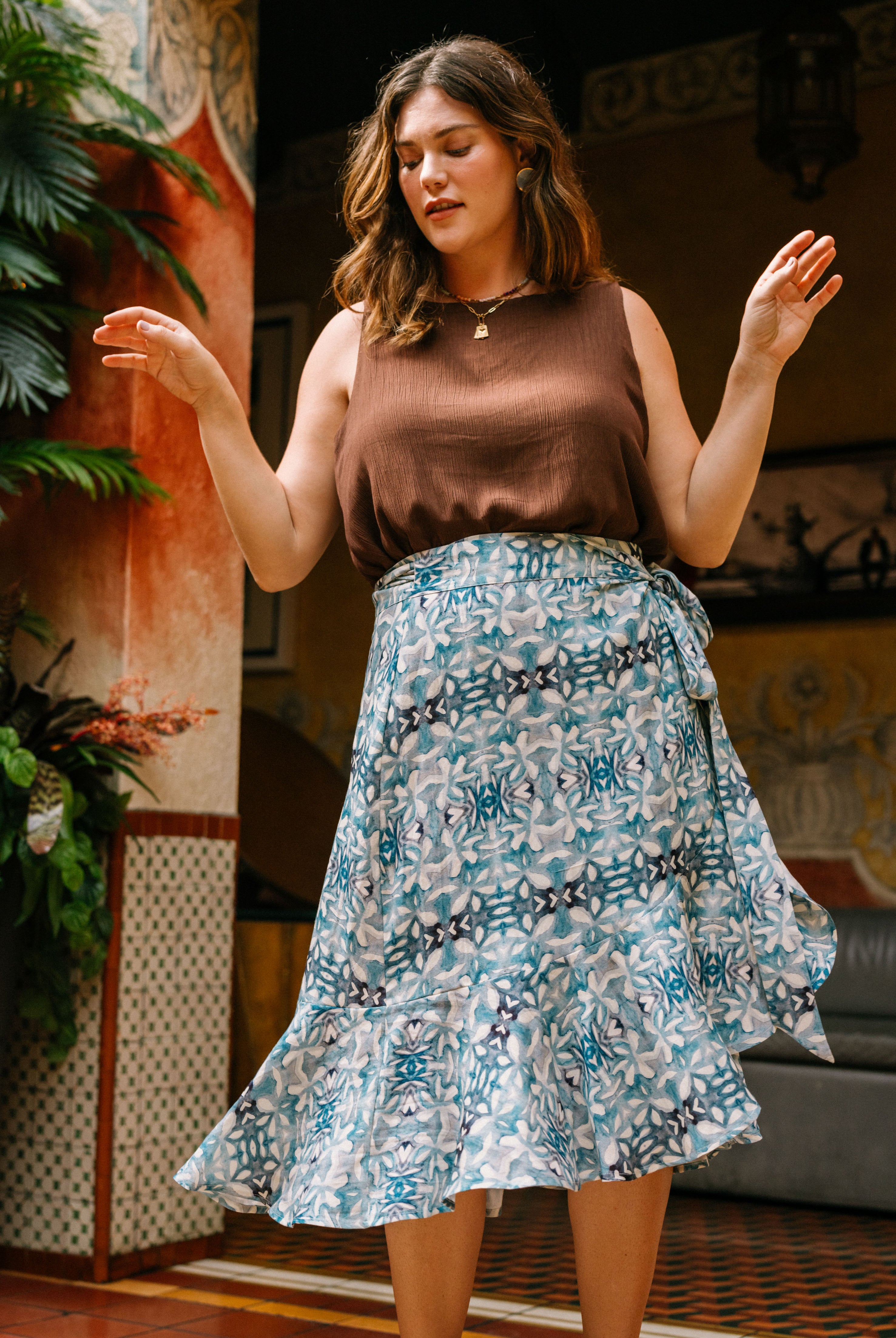 The Ruffle Skirt is a flowy skirt that hits mid-calf. It is a wrap skirt made out of blue and white fabric with a geometric, mediterannean-inspired print. The skirt wraps around and is fastened at the hip with a fabric tie in the same fabric as the skirt. The skirt has a slight ruffled effect at the bottom.