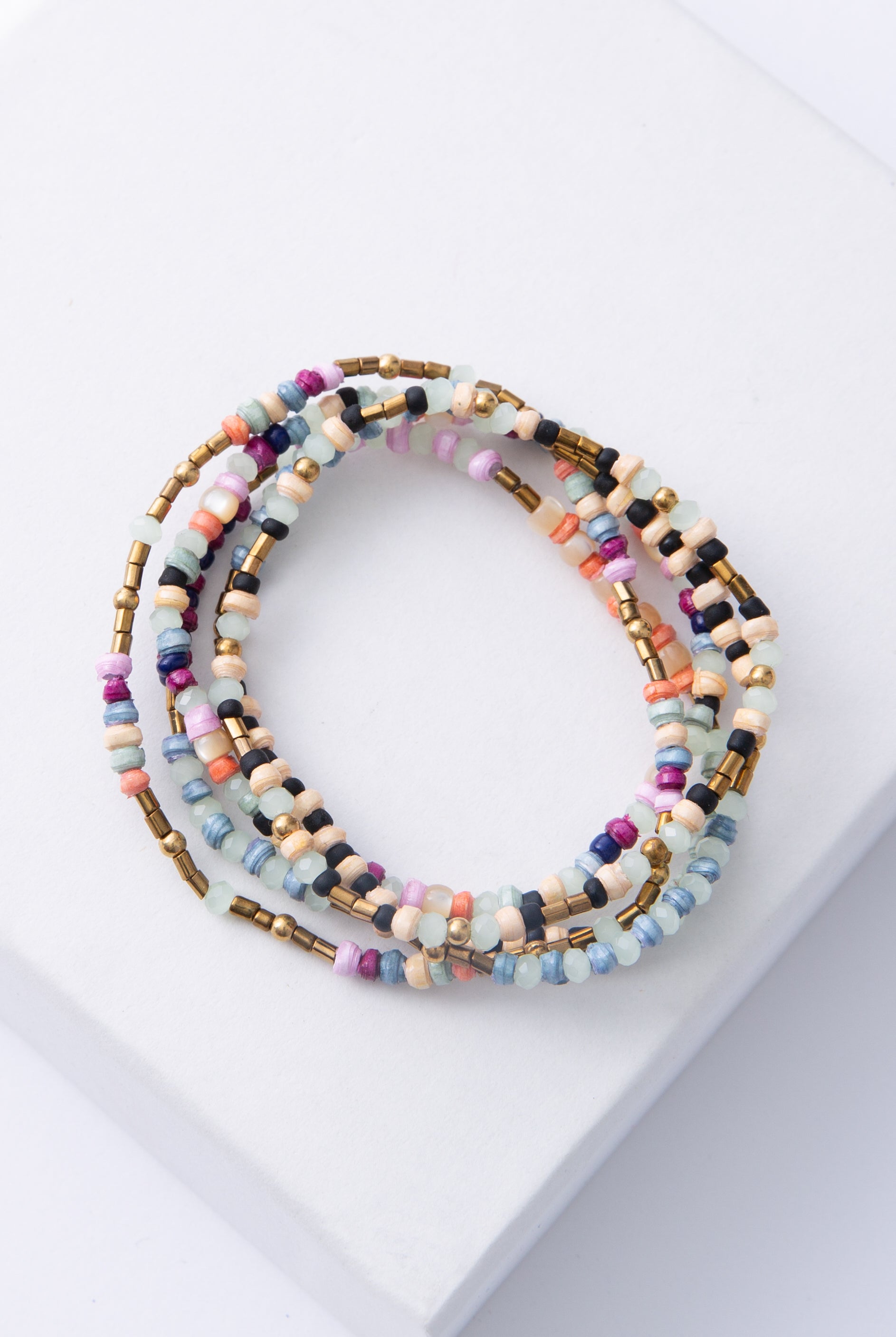 The Saltwater Stack includes five dainty beaded bracelets. Each bracelet features different colored paper beads in shades of cream, pink, blue, purple, and cream. The bracelets also feature mint-colored crystal beads, brass beads, and mother-of-pearl beads.