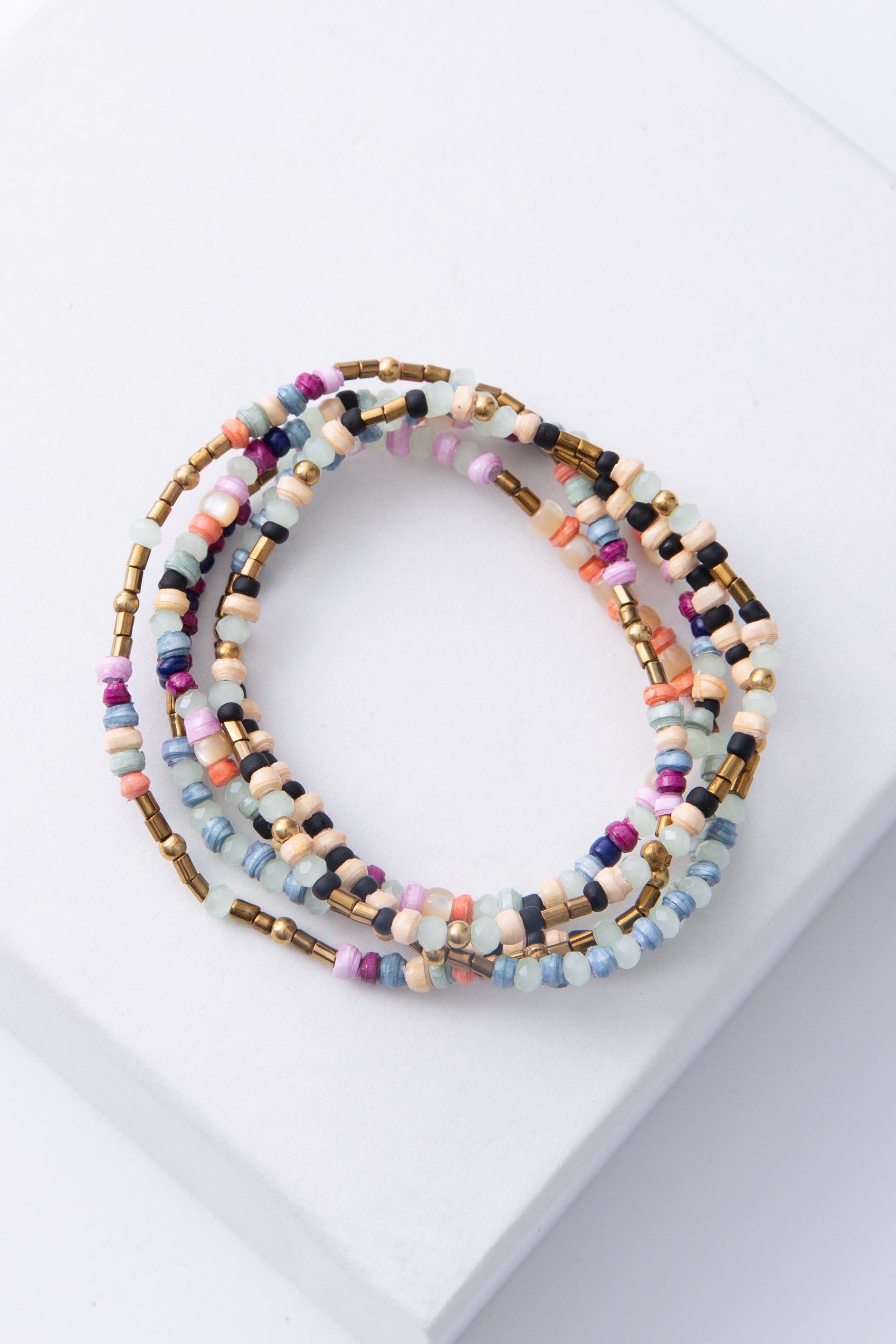 The Saltwater Stack includes five dainty beaded bracelets. Each bracelet features different colored paper beads in shades of cream, pink, blue, purple, and cream. The bracelets also feature mint-colored crystal beads, brass beads, and mother-of-pearl beads.