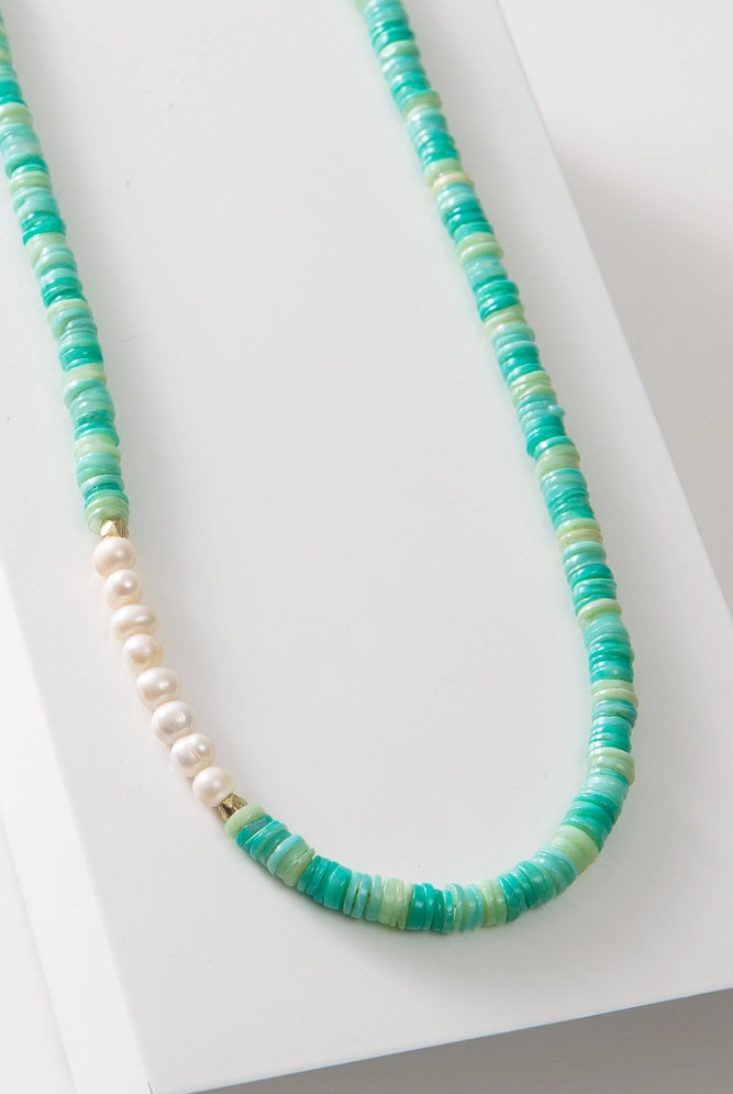 The Sea Pearl Necklace is a single stranded necklace composed of tiny disc-shaped shell beads in varying shades of turquoise. At one section of the necklace there is a row of eight round pearls with a faceted gold bead on each side.