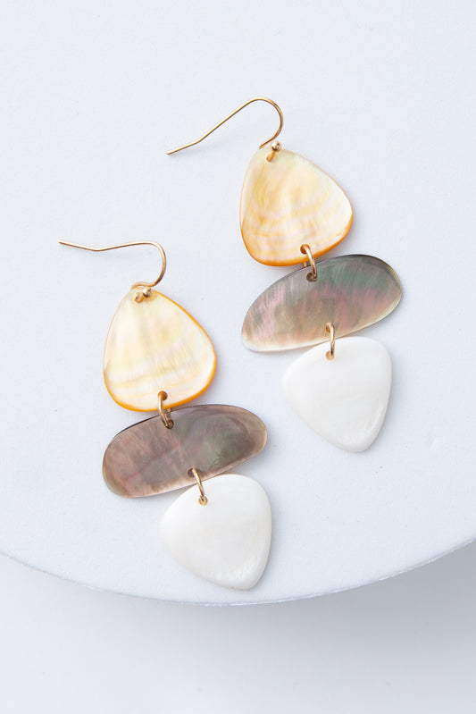 The Shell Stack Earrings are dangly earrings with brass ear hooks. Three flat pieces of shell are stacked on top of each other for a sculptural look. The top piece has caramel hues, the middle has grey hues, and the bottom has white hues. Each piece is connected by a brass ring.