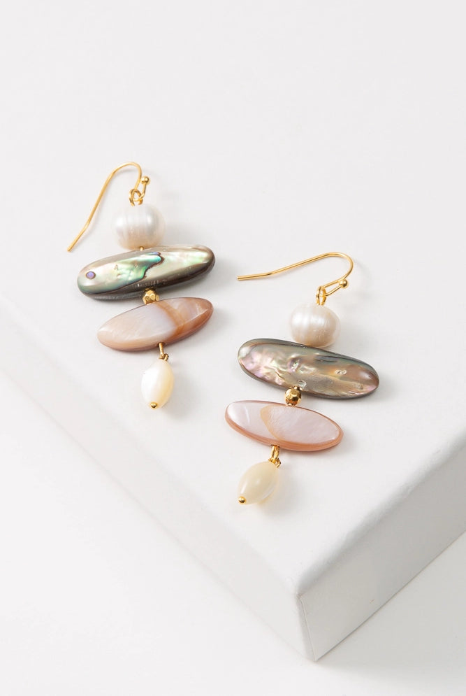 The Shell Trove Earrings are composed of gold ear hooks with pearl and shell pieces hanging down vertically, giving them a stacked look. At the top of the stack is a round pearl. Below this is an iridescent black shell piece, followed by a slightly narrower iridescent peach shell piece. At the bottom of the stack is an oval-shaped pearl bead.
