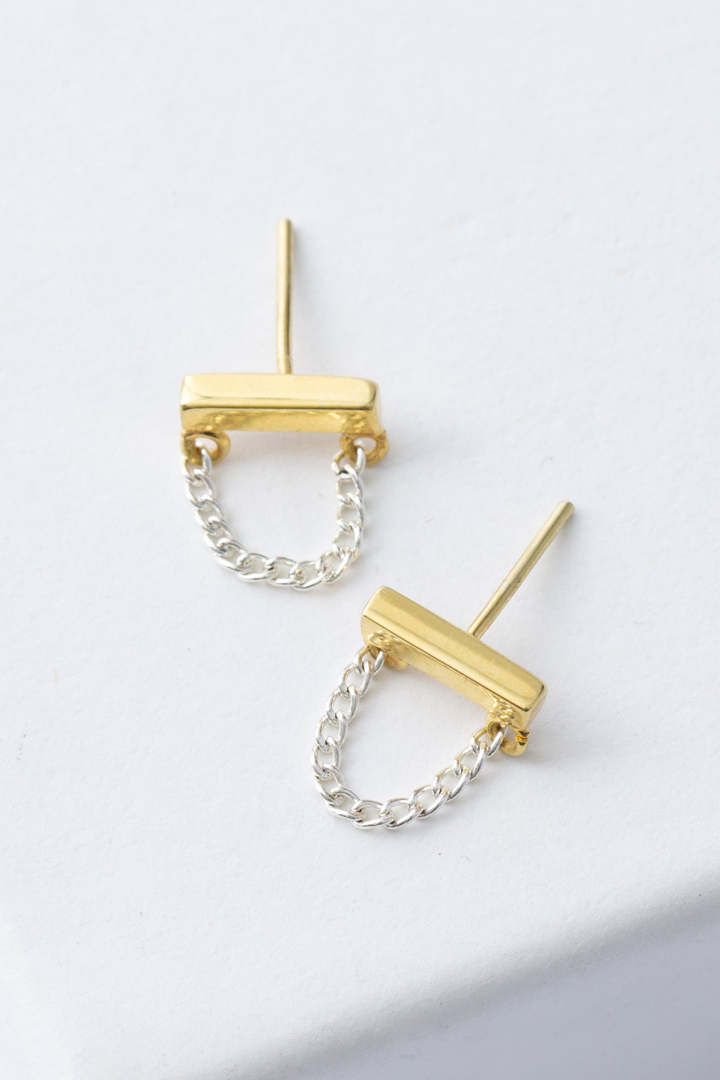 The Silver Lining Studs are silver and gold post earrings. A horizontal gold bar is accented with a silver chain that hangs down in a loop. 