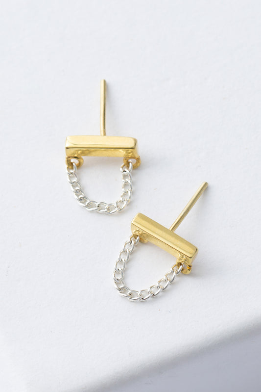 The Silver Lining Studs are silver and gold post earrings. A horizontal gold bar is accented with a silver chain that hangs down in a loop. 