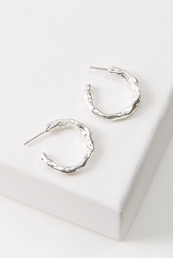 The Molten Metal Hoops are midi-sized hoops composed entirely of shiny silver metal. The metal has a rough, irregular shape that gives the earrings an organic feel. The hoops are open at the back.