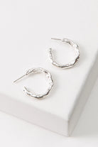 The Molten Metal Hoops are midi-sized hoops composed entirely of shiny silver metal. The metal has a rough, irregular shape that gives the earrings an organic feel. The hoops are open at the back.