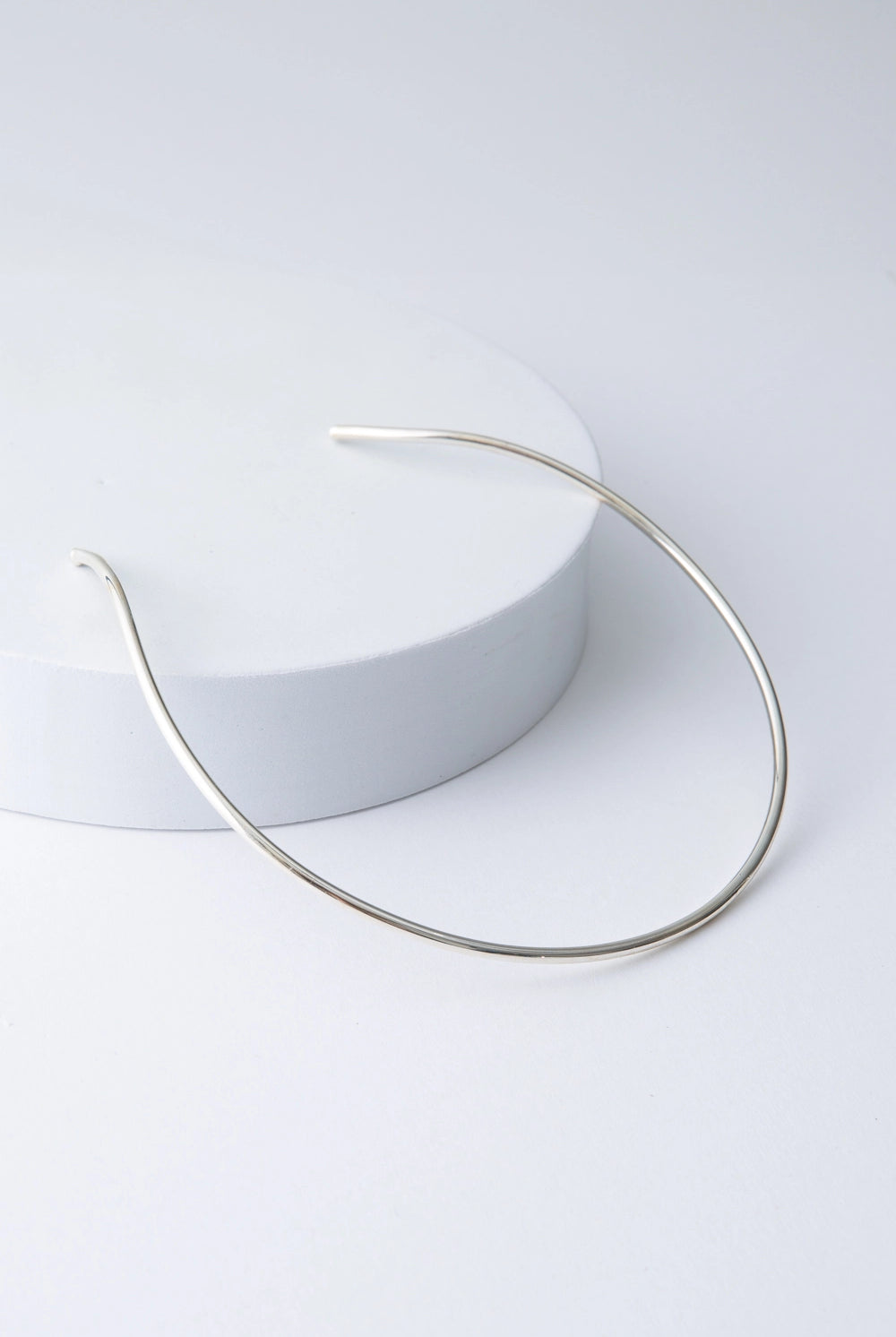 This is a choker-style necklace composed of a single bar of shiny silver plated bronze that forms a semi-circle. It is open in the back, allowing it to be slid onto the neck.