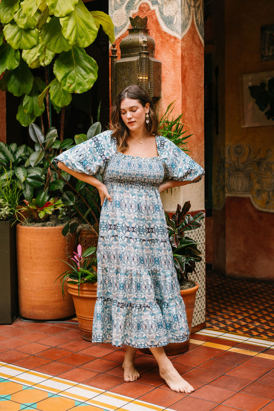 The Smocked Maxi Dress is a flowy dress with short, roomy sleeves. The long dress hits roughly at the ankle. It is made out of blue and white fabric with a geometric, mediterannean-inspired print. The chest area of the dress is stitched with elastic, making it stretchy. The sleeves can be worn up on the shoulders or pulled down on the sides of the shoulders. 