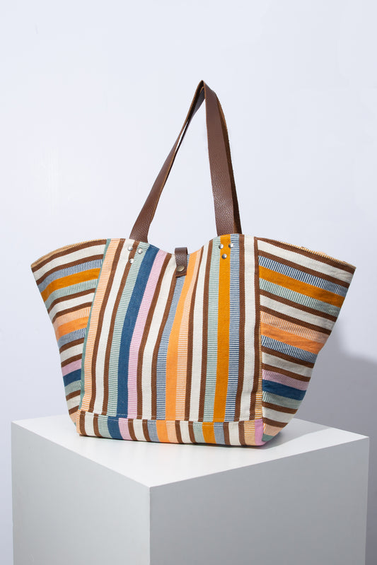 The Sunkissed Tote is a sturdy fabric tote composed of cotton fabric with vertical stripes in shades of blue, pink, maroon, mint, and orange. The bag has a flat base that allows it to sit upright. It is narrower at the base and then tapers out towards the top, making it wider at the top than it is at the bottom. It has a small leather snap closure and two brown leather handles. 
