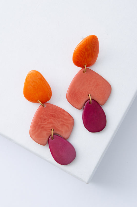 The Sorbet Sunset Earrings are chandelier-style earrings composed of pieces of flat, polished tagua seed stacked on top of each other. Attached to the post is an orange, tear-drop shaped tagua piece. Below this is a coral tagua piece shaped like an upside-down V. At the bottom hangs a maroon teardrop piece. The three pieces are attached with brass rings.