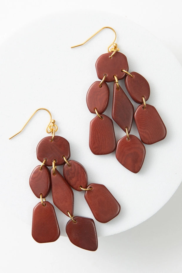 The Tagua Cascada Earrings are chandelier-style earrings with gold ear hooks. They are composed of polished pieces of tagua seed that have been cut into geometric shapes. The tagua is a rich rust shade. Each piece is linked with a gold jump ring to form a chandelier look.