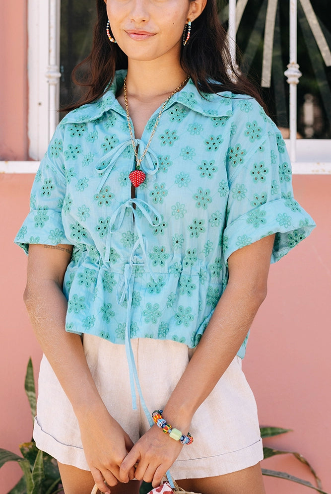 The Tie Cinch Blouse is a short-sleeve top composed of blue cotton fabric covered in a floral eyelet pattern. There is a drawstring at the waist that can be tightened and tied, creating a ruffled look below it. The top has a collar and is open down the middle, with two bow tie closures.