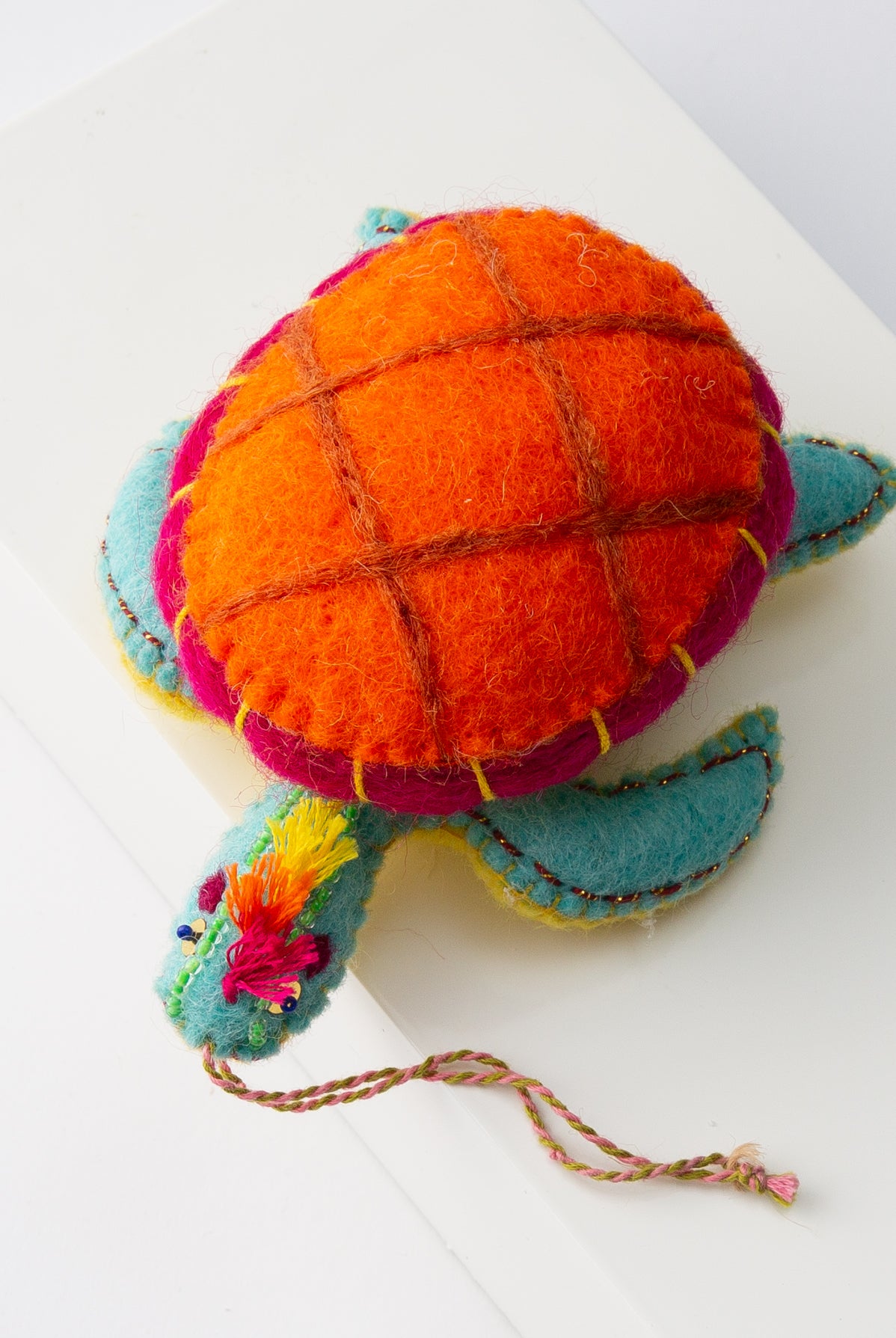 The Tiptop Turtle Ornament is shown from overhead. It is a small stuffed turtle made of fabric. The shell is bright orange and hot pink. The turtle is teal with a yellow belly. Festive thread fringe sticks out from the top of the turtle's head. A braided string attached to the body allows the ornament to be hung.