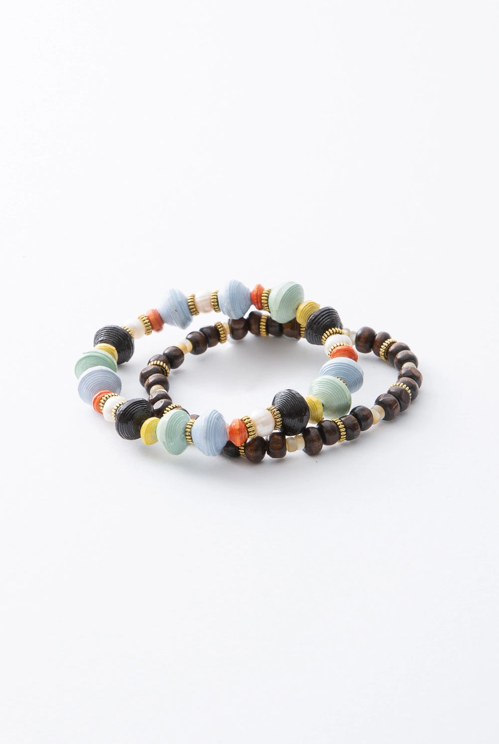 The two bracelets included in the Ubuntu set are shown stacked. Both are stretchy, single-strand beaded bracelets. One bracelet is strung with hand rolled paper beads in shades of light blue, mint, orange, yellow, and black, along with decorative brass beads. The other bracelet is strung with dark brown ox horn beads and fresh water pearl beads, accented with the same decorative brass beads.