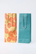 Two examples of the Upcycled Metallic Wine Bags are pictured. Both are tall, skinny, and rectangular. Each has two woven cord handles in a complementary color attached to the top of the bag. The bags are each a different color and pattern, but both have metallic elements that give the paper a shiny look.