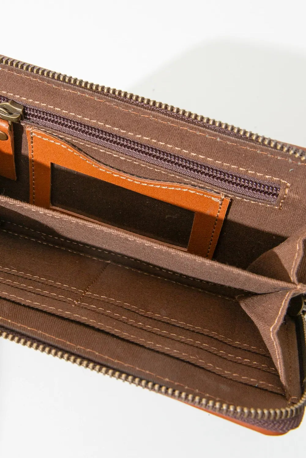 The Vaquera Wallet is shown unzipped. One interior wall has a zip pocket, as well as an ID slot pocket that is clear in the center and outlined in orange leather. There is a divider in the center of the wallet, dividing it into two main compartments. The opposite wall has several fabric slot pockets for cards.