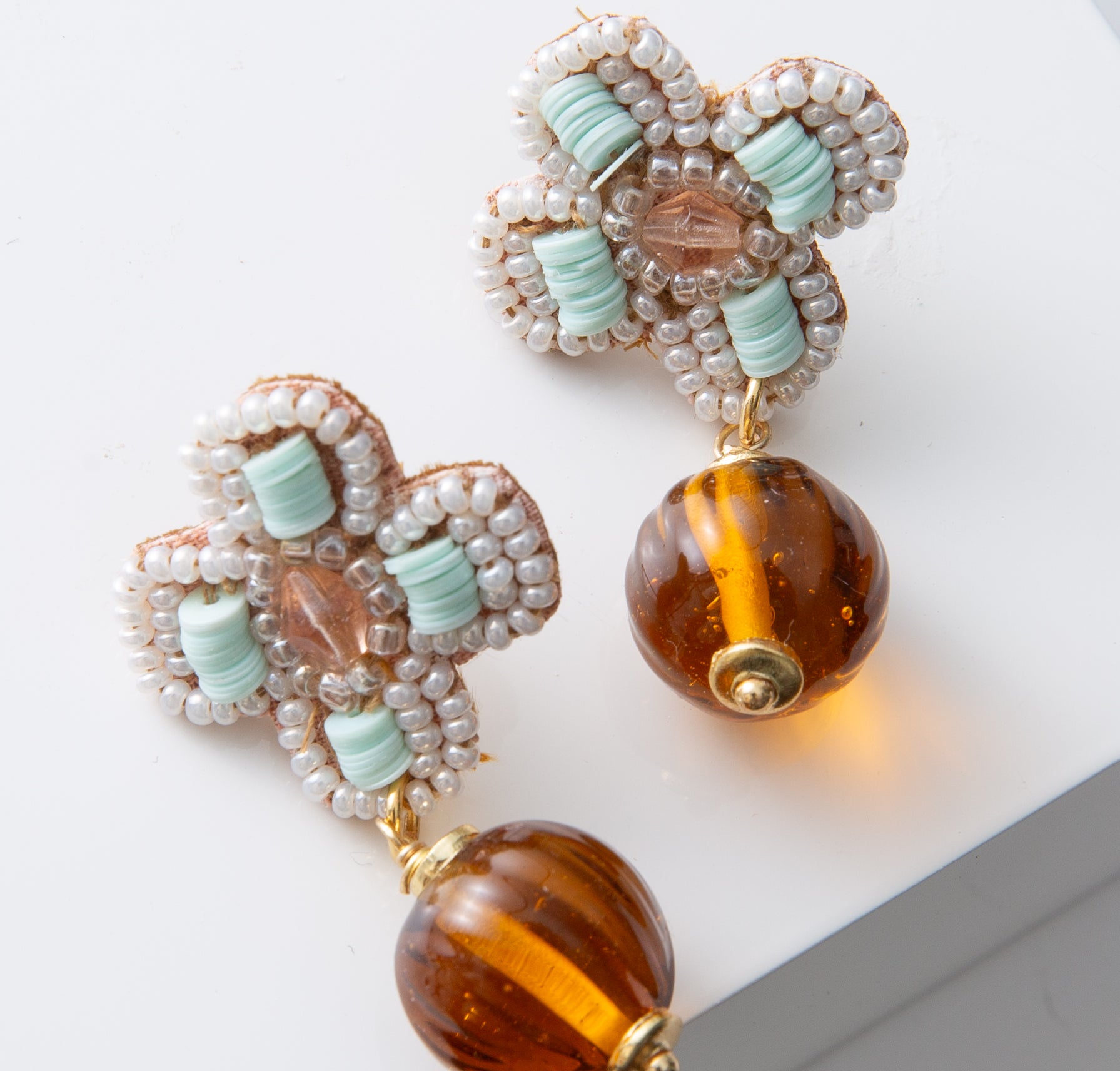 The post of the Vintage Amber Drop Earrings are adorned with mint, blush, and white beads forming the shape of a four-petaled flower. Hanging down from the flower is a spherical amber glass bead with a ribbed texture.