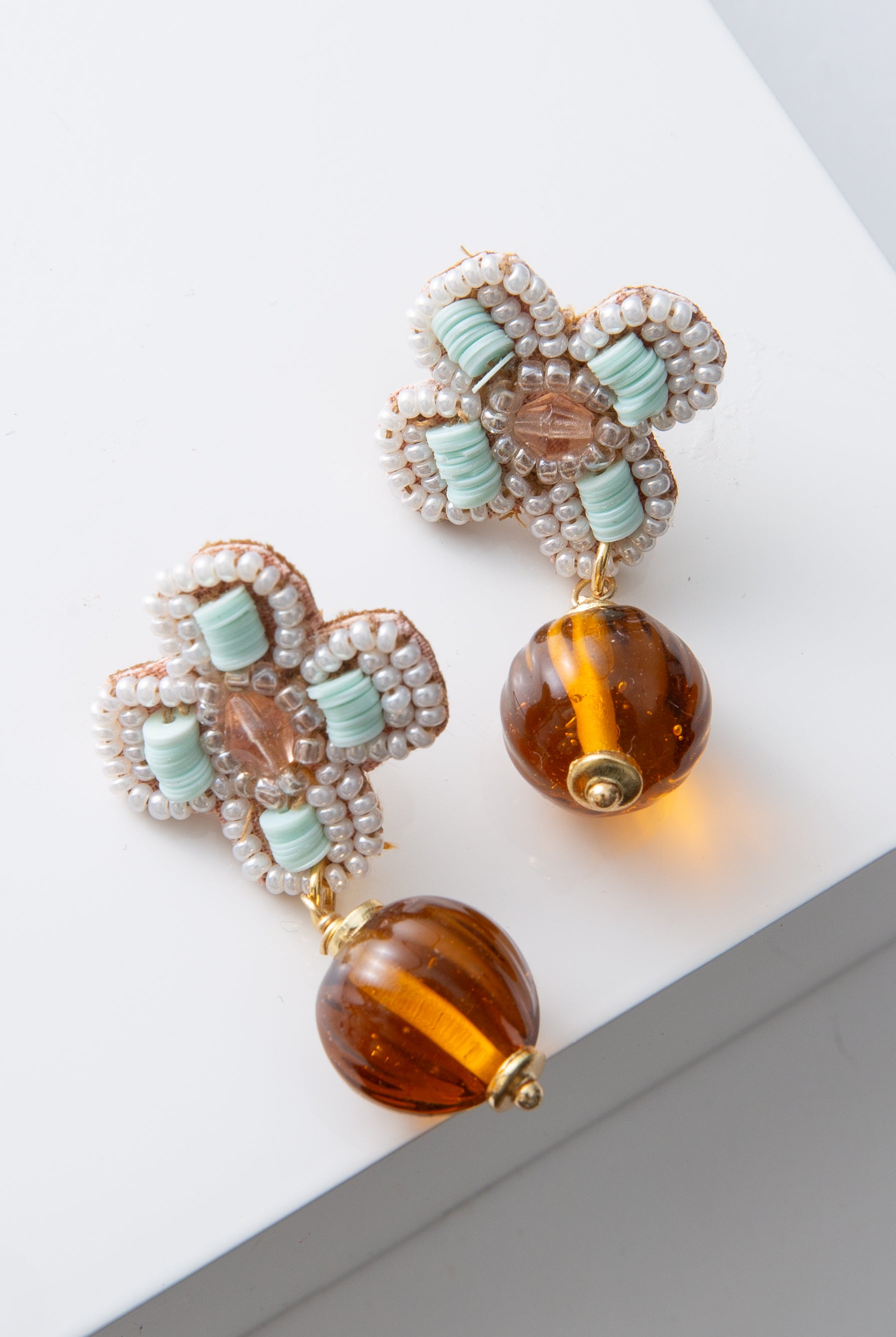 The post of the Vintage Amber Drop Earrings are adorned with mint, blush, and white beads forming the shape of a four-petaled flower. Hanging down from the flower is a spherical amber glass bead with a ribbed texture.