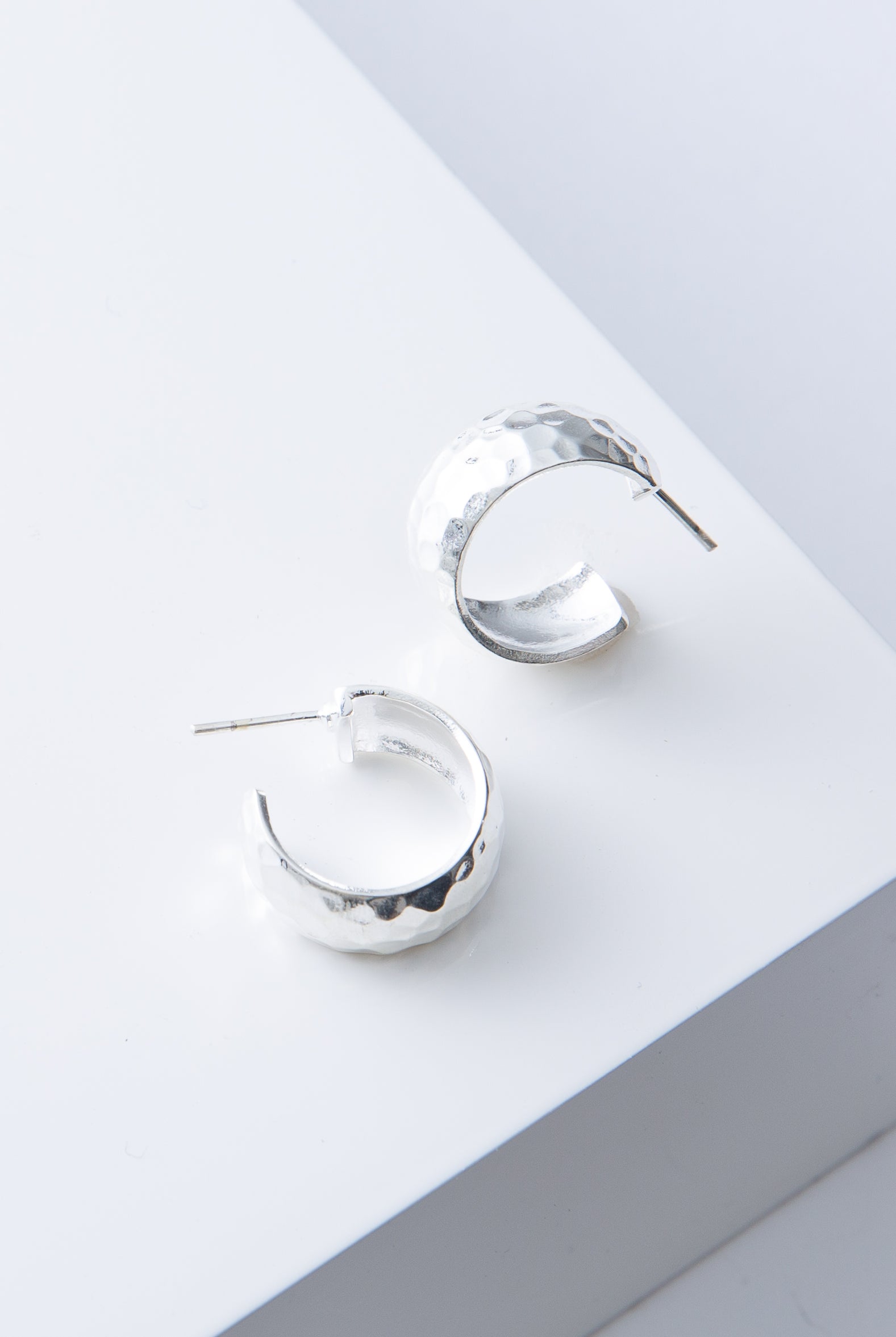 These are chunky, demi-hoop earrings that are open at the back. They are composed of flattened silver plated bronze with a hammered texture. The hoops are attached to a steel post.