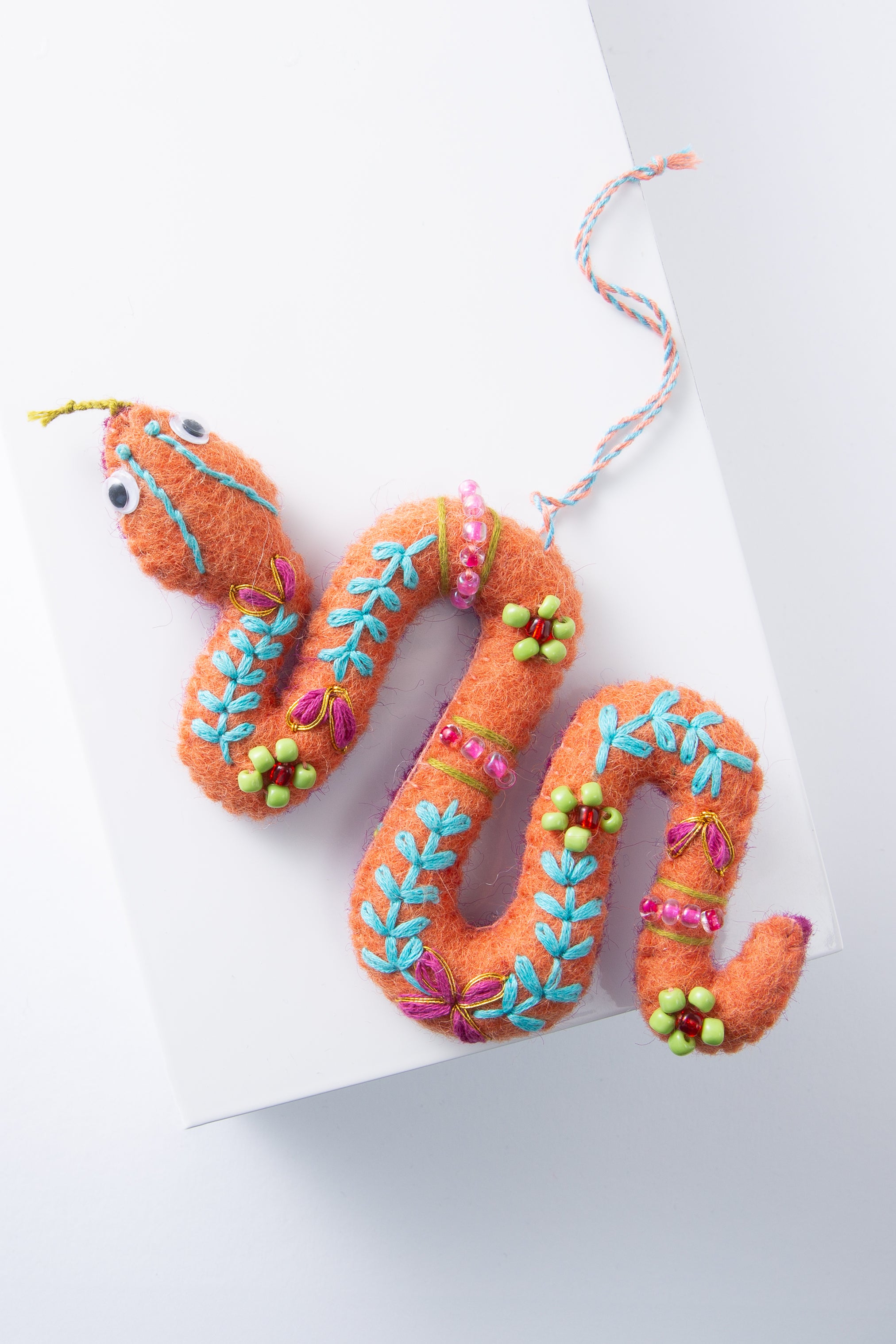 The Squiggly Snake Ornament is composed of brightly colored pieces of wool stitched together to form a three dimensional coiled snake. The body is orange, and there is aqua and pink embroidery adorning the body. Green glass beads are also stitched into floral decorations along the body. The ornament hangs from a loop composed of twisted blue and orange threads.