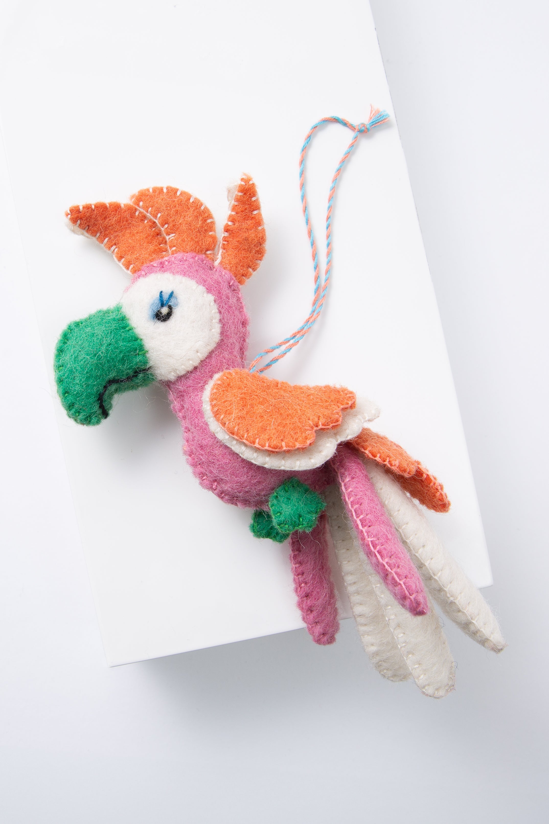The Coo Coo Cockatoo Ornament is composed of brightly colored pieces of wool stitched together to form a three dimensional bird. The body is pink, the beak is green, and the feathers are orange, white, and pink. There is an eye and eyelashes stitched onto the face. The ornament hangs from a loop composed of twisted blue and orange threads.