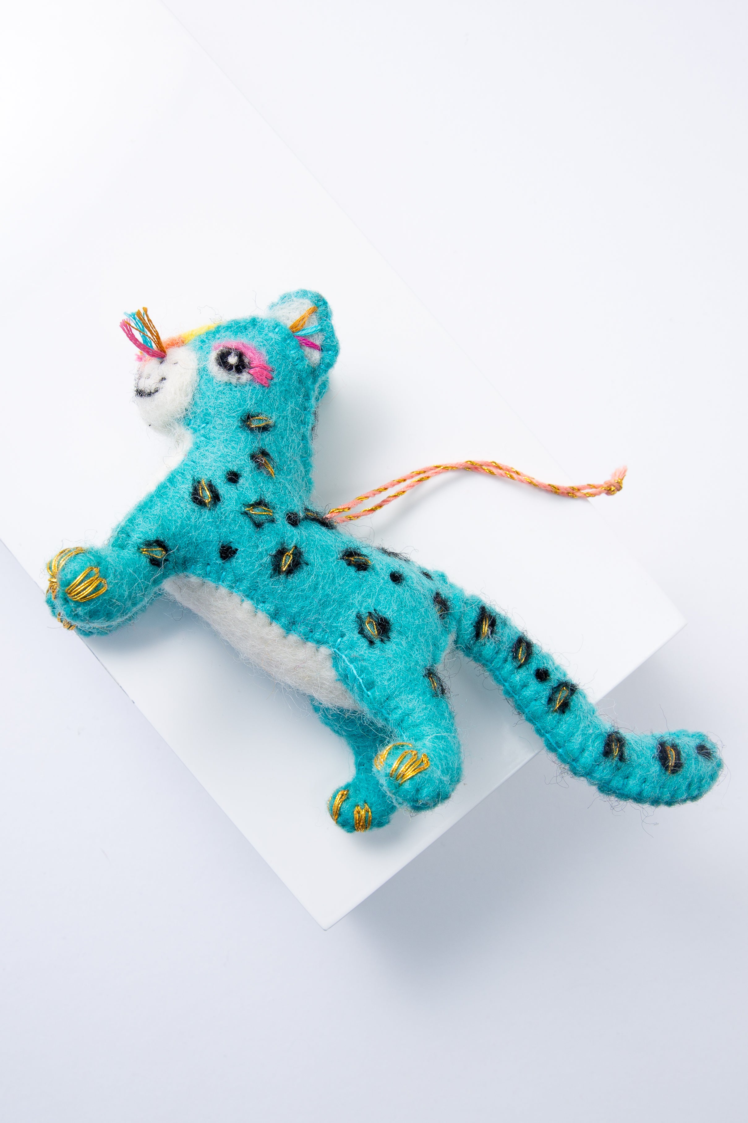 The Lucky Leopard Ornament is composed of brightly colored pieces of wool stitched together to form a three dimensional leopard. The body is turquoise, and the snout, belly, and inner ears are white. There is an eye and eyelashes stitched onto the face, and black and gold spots stitched all over the body. The ornament hangs from a loop composed of orange thread.
