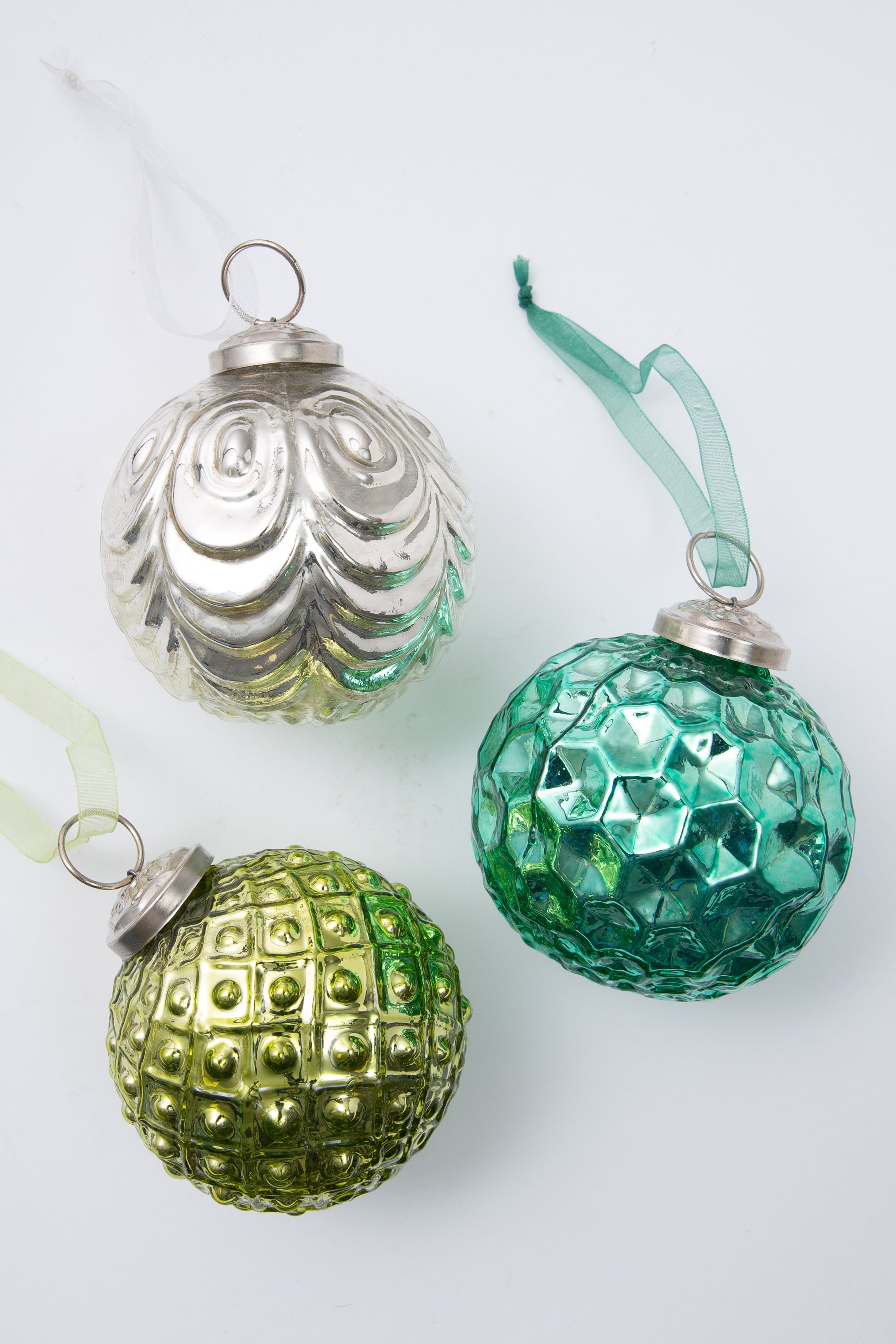 The three ornaments included in the Metallic Glass Ornament Set are shown. All three are round bauble ornaments made of glass with a metallic finish. Each has a different textured pattern and color. One is silver, one is aqua, and one is light green. Each ornament has silver rings with a complementary colored ribbon attached.