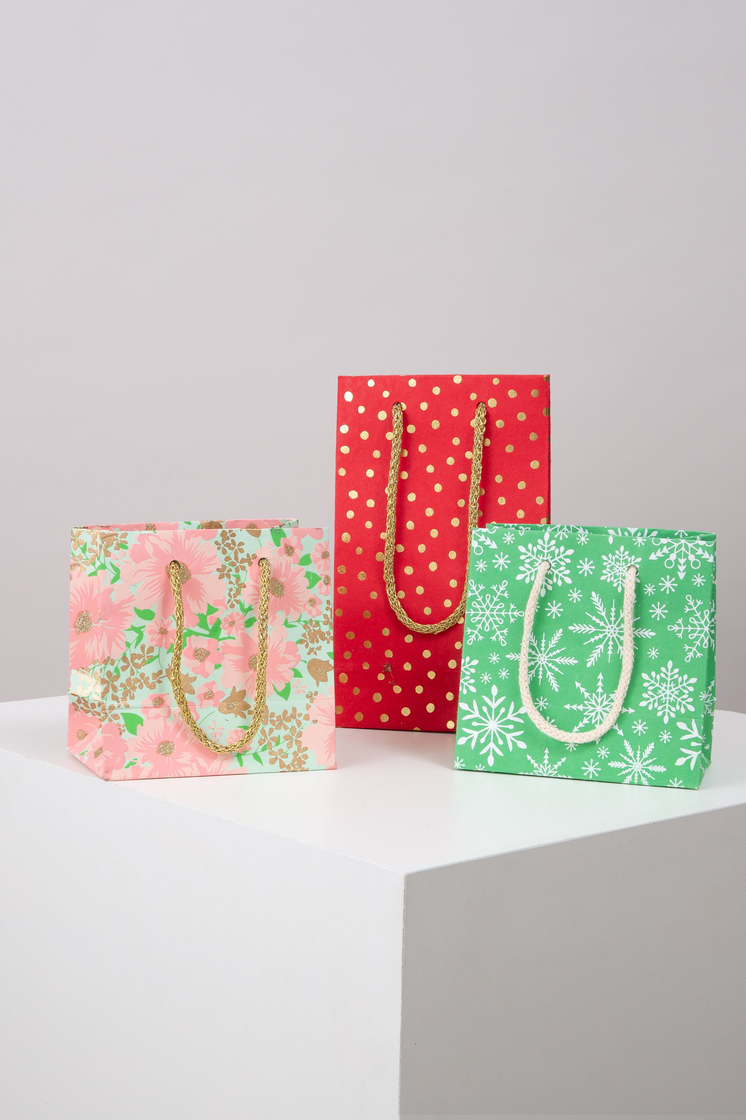 Three examples of the Upcycled Gift Bags are pictured. Two are square, and one is tall and skinny. Each has two woven cord handles in a complementary color attached to the top of the bag. The bags are each a different color and pattern. One has a floral pattern, one is polka dotted, and one has a snowflake pattern.