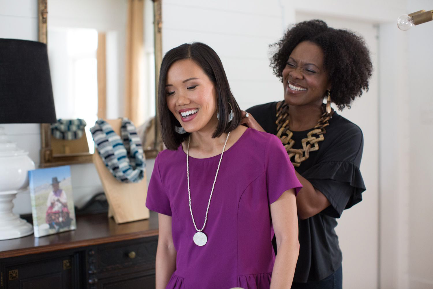 two women smiling, one is putting a necklace on the other. 